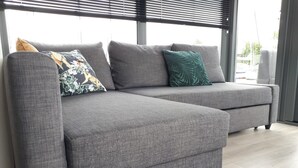 Un sofá seccional gris con un cojín estampado con flores y un cojín con diseño de hojas, ubicado frente a una ventana con persianas.