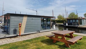 Una estructura flotante moderna con fachada de vidrio, una mesa de picnic de madera y un área verde frente a ella.