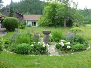 Un jardín con una fuente de piedra, rodeado de diversas flores y arbustos, y una casa al fondo.