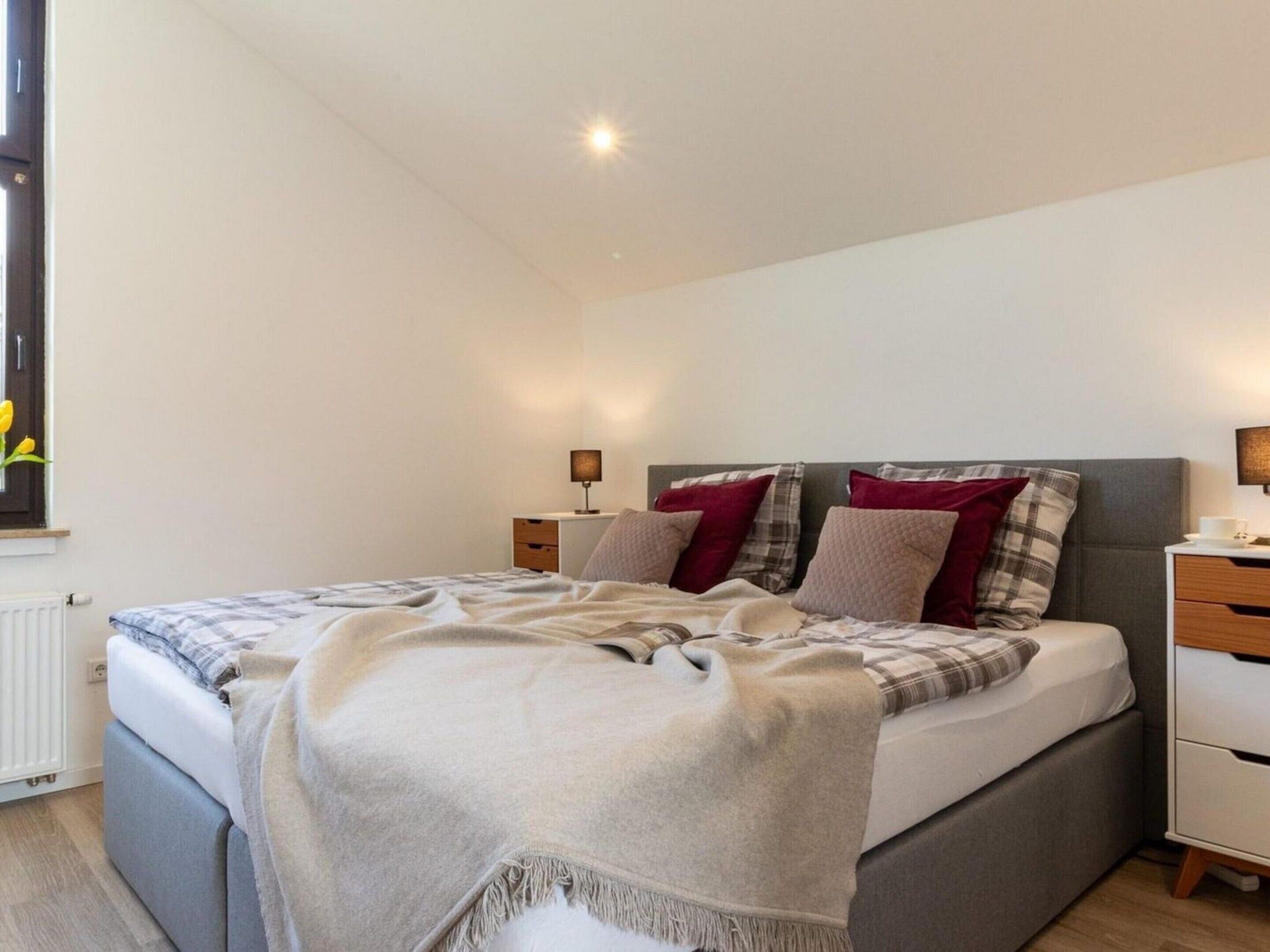 A modern bedroom with a large bed, bedside tables, a window with flowers, and a radiator.
