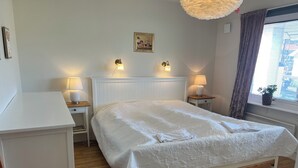 A neatly arranged bedroom with a large bed, two bedside tables with lamps, a wooden nightstand, a white dresser, and a window with curtains.