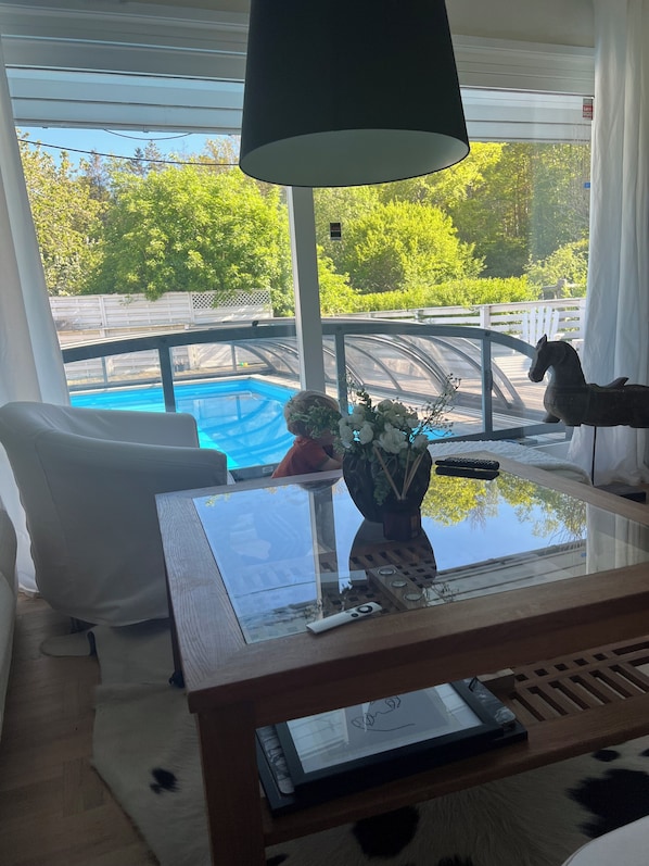 Una habitación con una mesa de vidrio, una silla blanca, una lámpara, un florero con flores y un control remoto, con una piscina visible a través de la ventana.