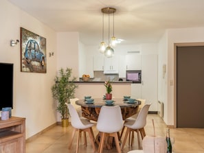 A modern dining area with a round table, white chairs, a potted plant, and a kitchen in the background.