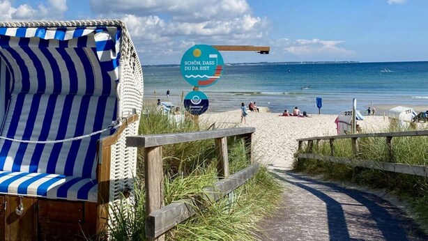 En träkonstruktion vid stranden med ett randigt solskydd, en skylt med texten ”Schön, Dass du bist” (Vackert, du är vacker) och en gångväg som leder ner till stranden.