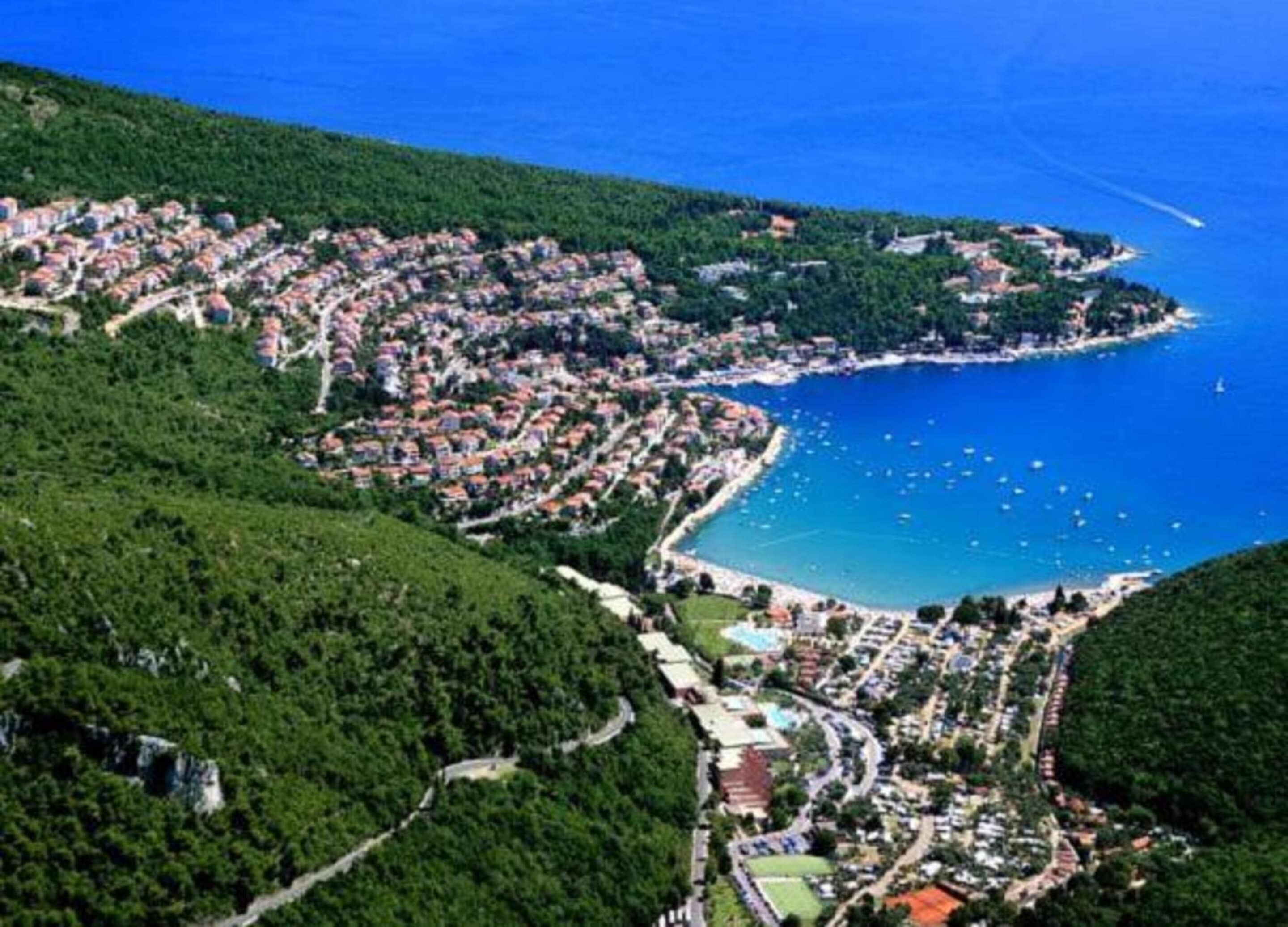 An aerial view of a coastal town with a dense cluster of buildings, a clear blue sea, and a winding road leading to the beach.