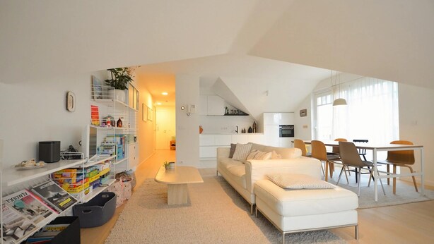 A modern living room with a white sofa, a coffee table, and a dining area with chairs and a table. There is a bookshelf with books and decorative items.