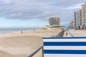 Una spiaggia con un edificio moderno caratteristico sullo sfondo, un percorso pedonale e alti edifici residenziali.