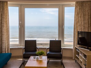 A room with a large window overlooking the sea, two chairs, a small table with a plant, and a TV on a stand. - Apartment With 1 Bedroom and Direct sea View (Middelkerke)