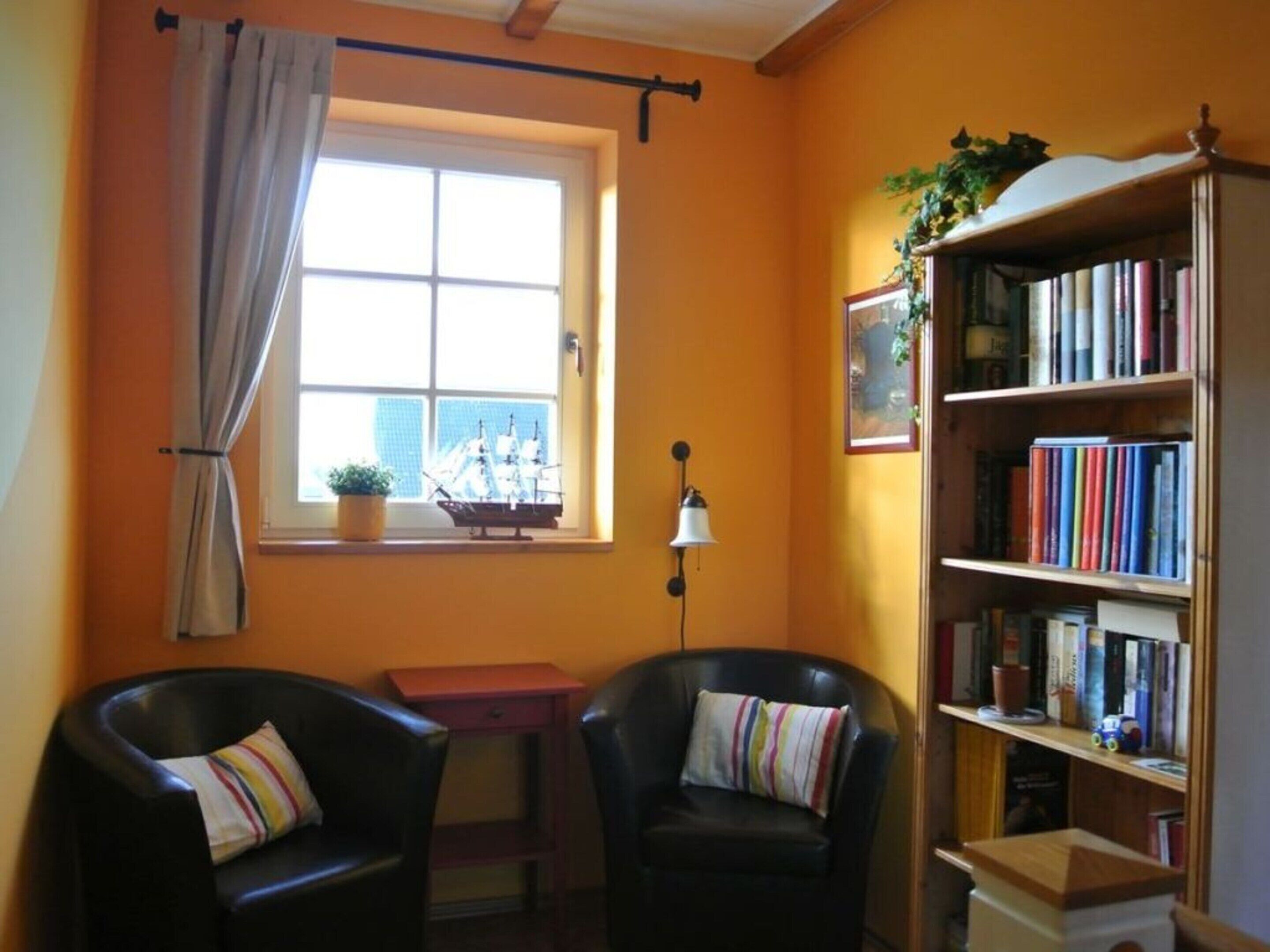 A room with two black leather chairs, a wooden bookshelf filled with books, a small table with a potted plant, and a window with a curtain.