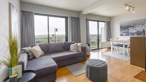 Un salon moderne avec un canapé gris, un petit pouf rond, une table à manger avec des chaises et de grandes fenêtres donnant sur la ville.