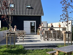 An outdoor patio area with wooden tables and benches, a black building with a tiled roof, and a clear blue sky. - Luxurious Studio on the Border of Zeeland (Stellendam)