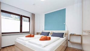 A double bed with two rolled towels, a bedside table, a nightstand, a window with blinds, and a blue accent wall. - Tranquil Apartment in Zell-kaimt (Zell (Mosel))