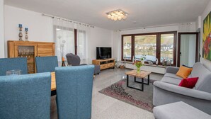 A modern living room with a sofa, blue chairs, a coffee table, a TV, and a large window with a view. - Tranquil Apartment in Zell-kaimt (Zell (Mosel))