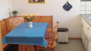 Ein kleiner Essbereich mit einem Holztisch und Stühlen, einer Tischdecke mit blauen Punkten, einer Vase mit Blumen, einer Uhr und einer Küchenzeile mit Schränken und einem Spülbecken.