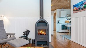 Una sala de estar acogedora con una estufa de leña, un sillón gris, una mesa de centro de madera con plantas, y una cocina al fondo.