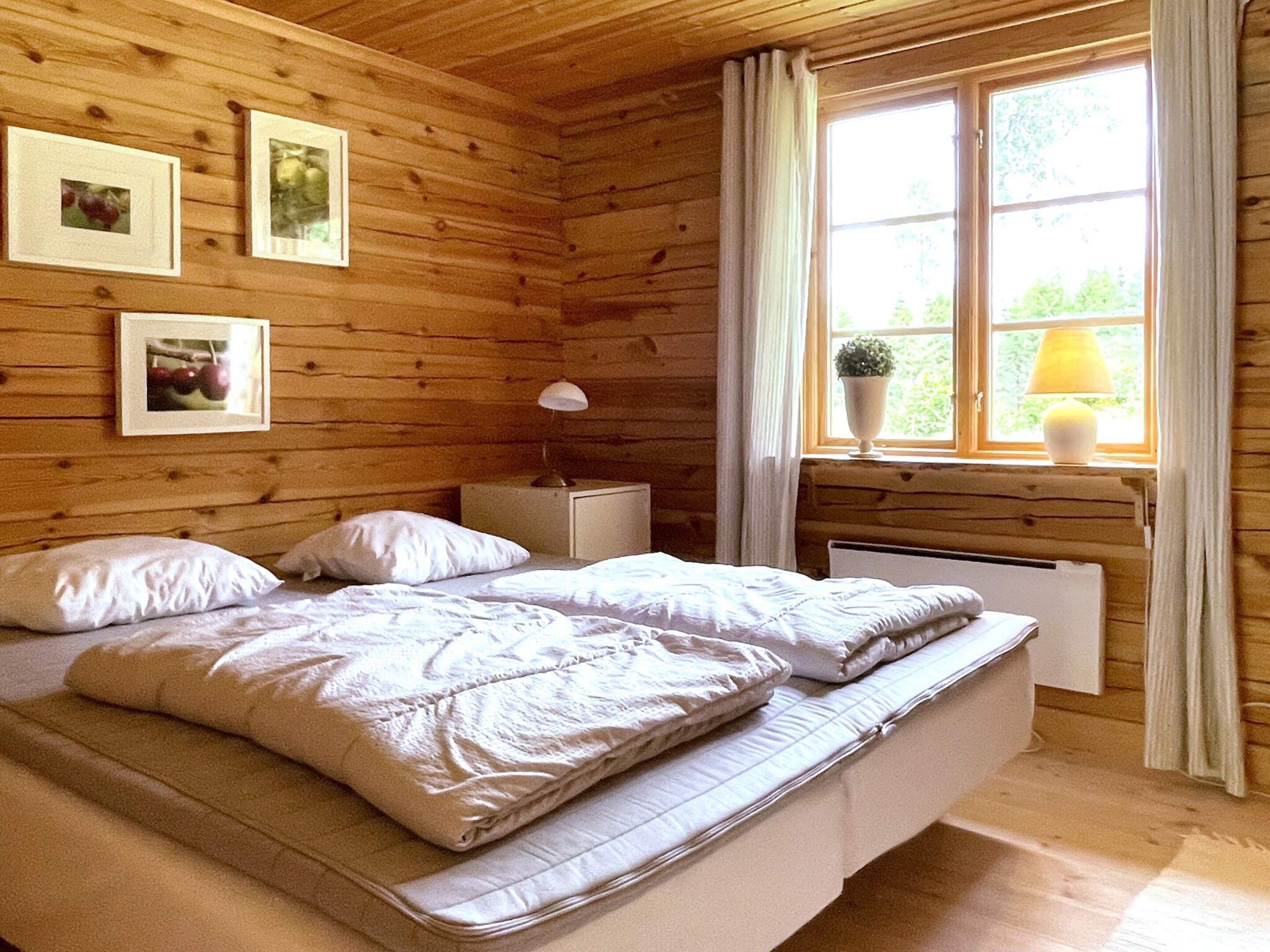 Un dormitorio de cabaña de madera con cama, mesita de noche, lámpara y ventana con cortinas.