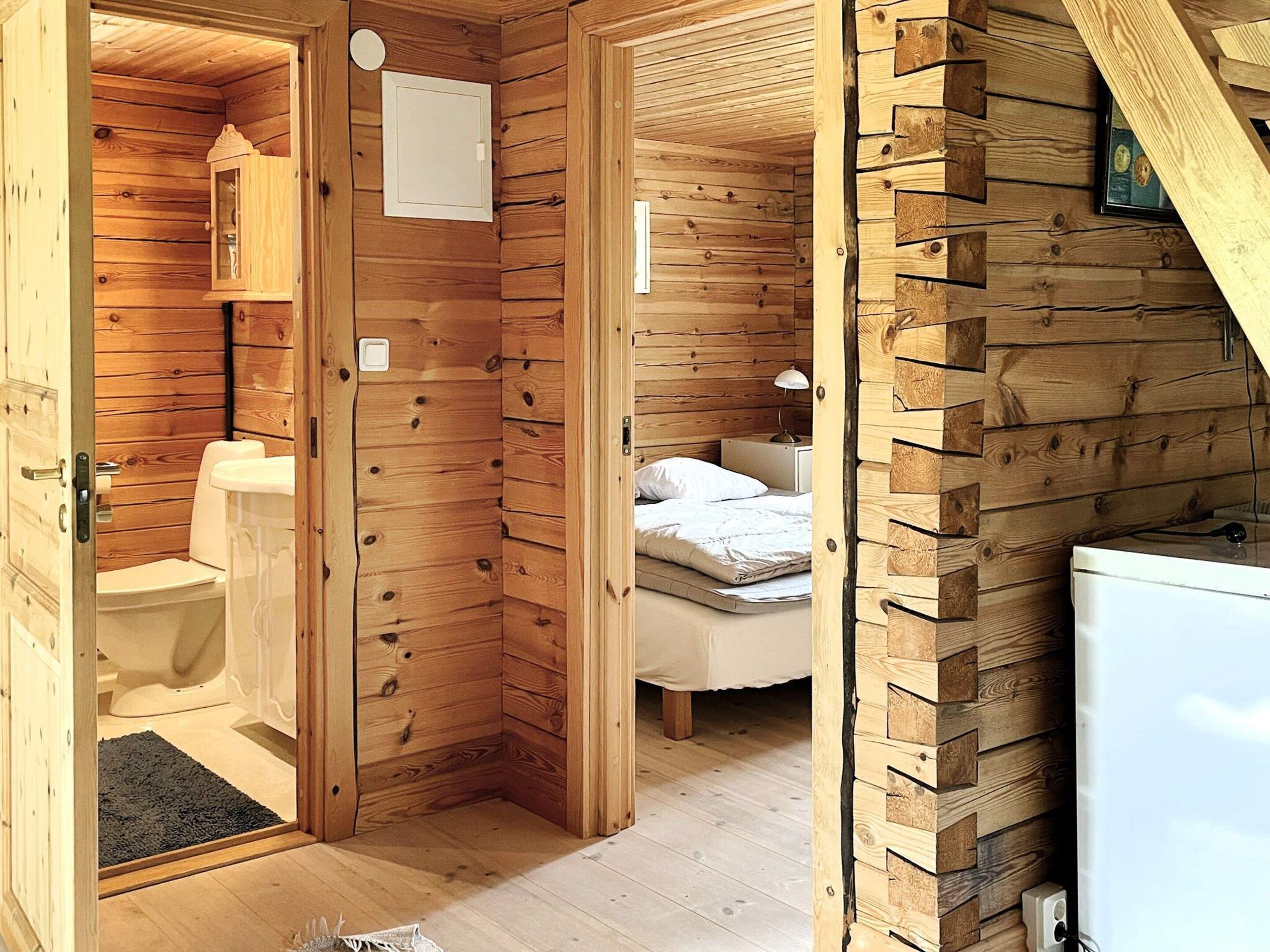 A wooden cabin interior with a bathroom, a bed, and a small kitchenette.