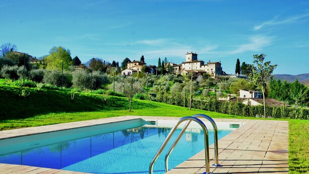 Une piscine à l’eau d’un bleu limpide, entourée d’une terrasse dallée. Au fond, on aperçoit des maisons sur une colline, sous un ciel dégagé.
