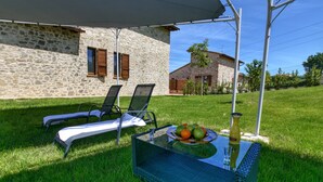 Un bâtiment en pierre avec une pelouse verdoyante, deux chaises longues et une table sur laquelle se trouvent des fruits et une bouteille.