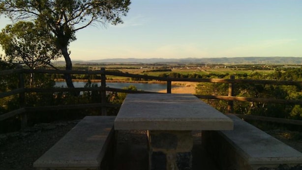 Una mesa de picnic con base de piedra, rodeada por una valla de madera, con vistas a un lago y colinas distantes.