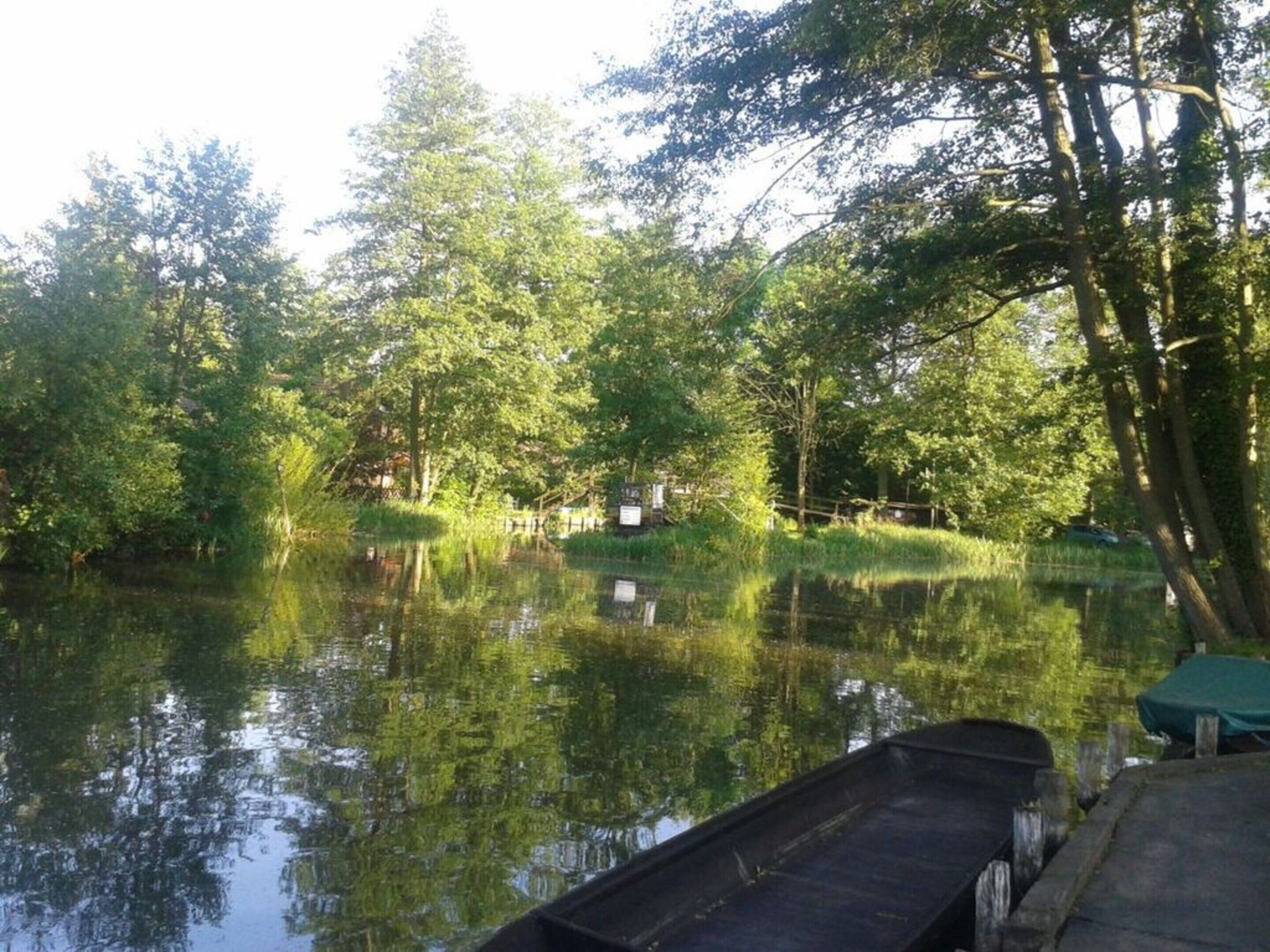 Tyyni joki, jonka pinnassa puut heijastuvat, laiturialue ja taustalla näkyvä opastekyltti.