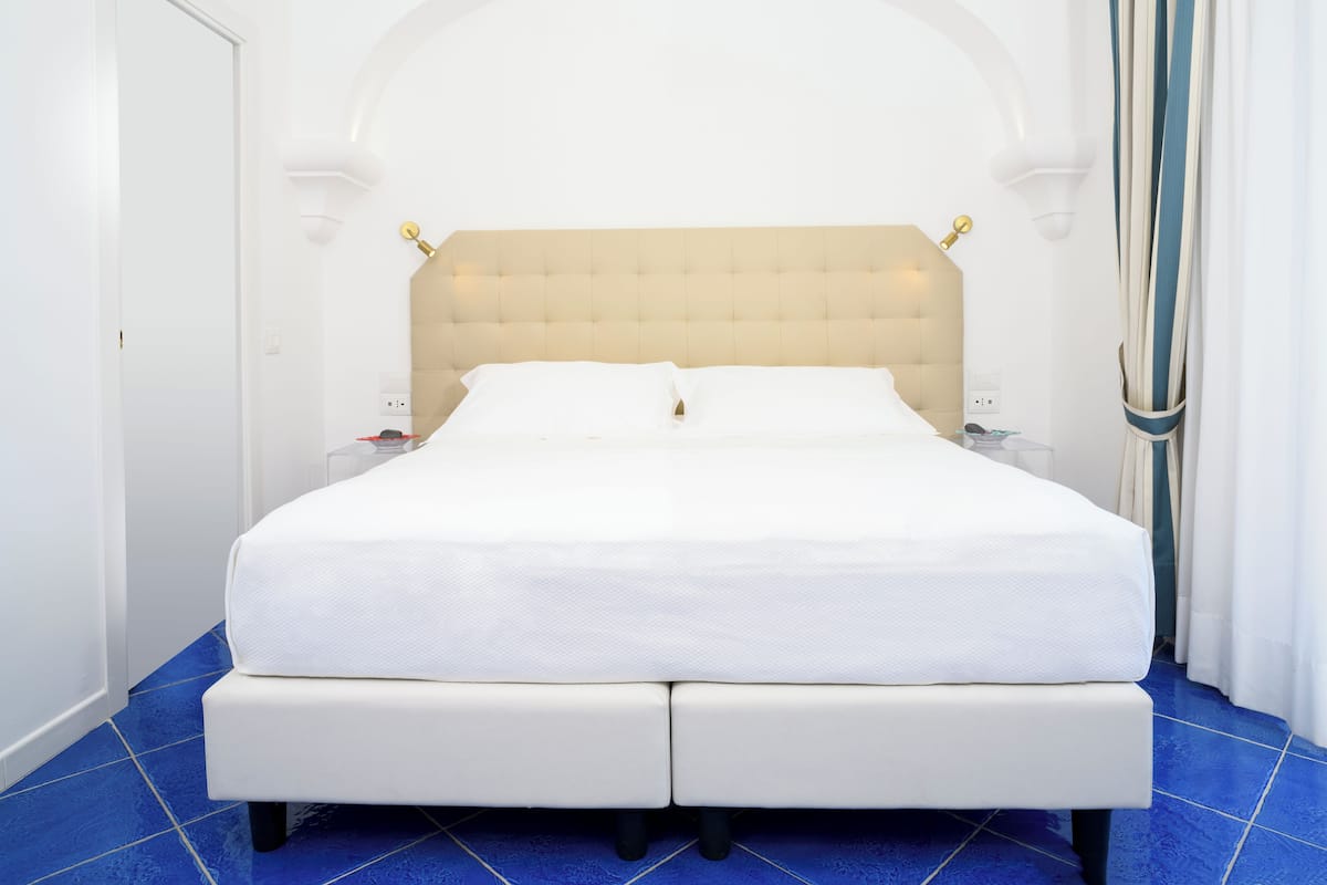 Un grand lit doté d’une tête de lit capitonnée, de draps blancs et d’une table de chevet avec une lampe.