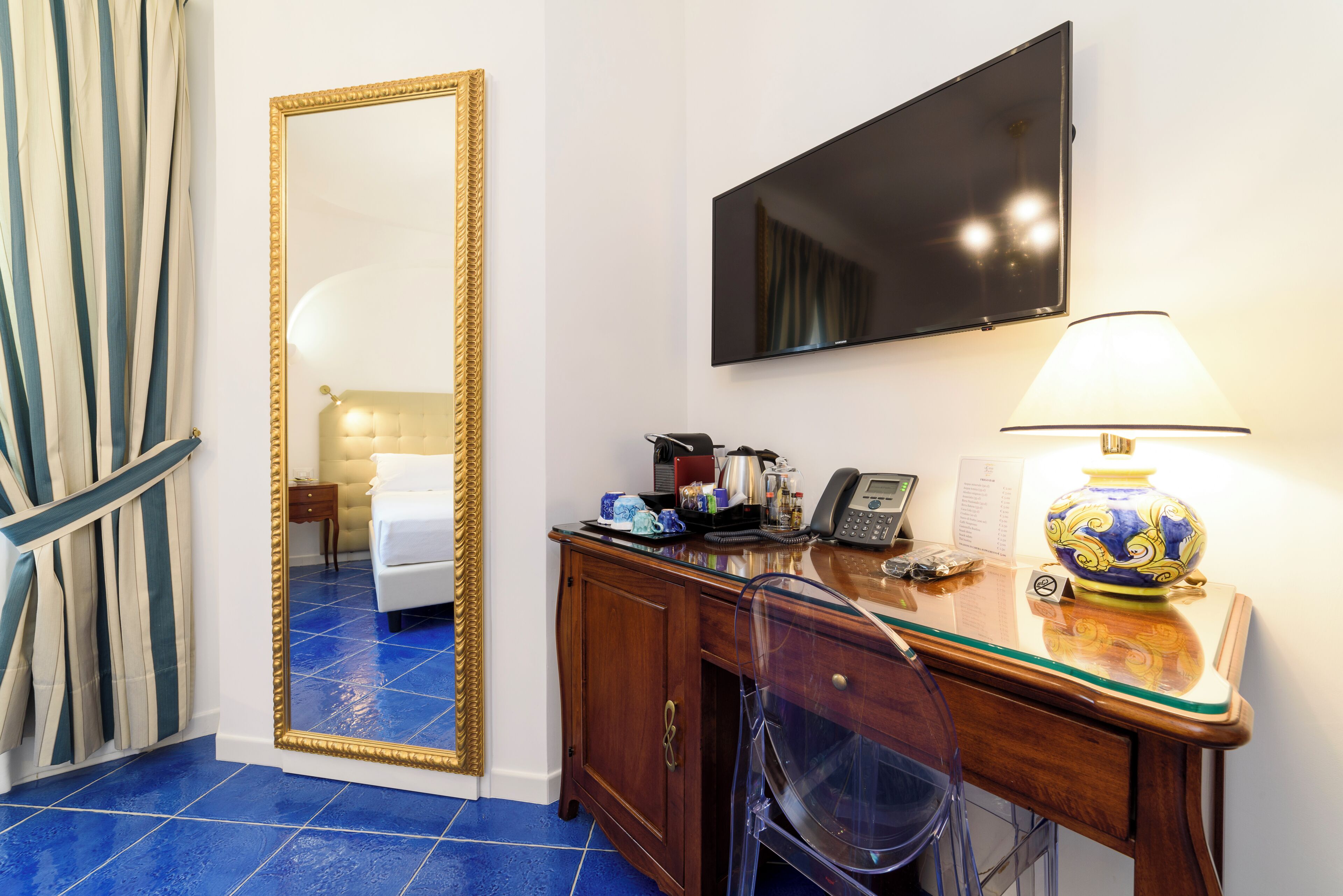 A hotel room with a large mirror, a flat-screen TV, a desk with a lamp, and a chair.