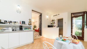 A modern kitchen with a white countertop, a coffee machine, and a variety of bottles. There is a dining area with a table set for two, featuring a plate of food and a cup. - La Casa di Paolo Boutique Hotel (Camerota)