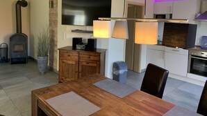 A modern living room with a wooden dining table, a fireplace, a TV, and a kitchen area with cabinets and appliances. - Just Before the Cape, Holiday Home B (Putgarten)