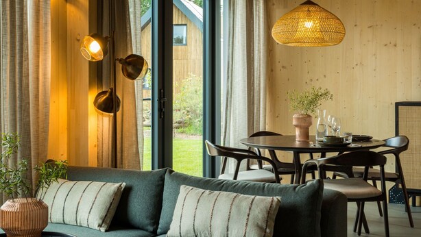 En hyggelig stue med en sofa, et spisebord med stole, en hængende lampe og udsigt til det fri gennem en glasdør.