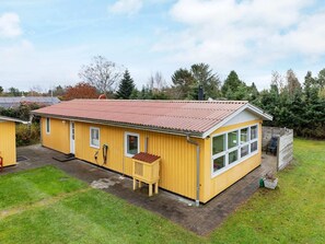 Ett gult, enplanshus med rött tak, en liten veranda och en inhägnad trädgård på baksidan.