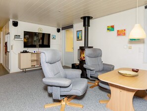 Une pièce avec deux fauteuils gris, une table en bois, un téléviseur, une cheminée et une horloge au mur.