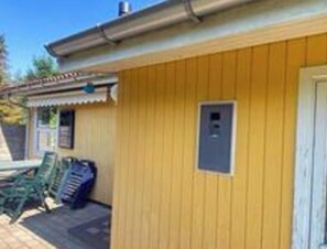 A yellow building with a door and a small window, a covered outdoor area with chairs, and a visible electrical box.