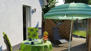 Eine Terrasse mit Tisch, der mit einer grünen Tischdecke bedeckt ist, zwei Stühlen, einem kleinen Blumentopf und einem gestreiften Sonnenschirm.