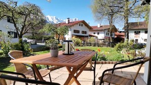 Un patio al aire libre con una mesa de madera y sillas de mimbre, rodeado por un jardín con diversas plantas y flores, y vistas a una cadena montañosa al fondo.