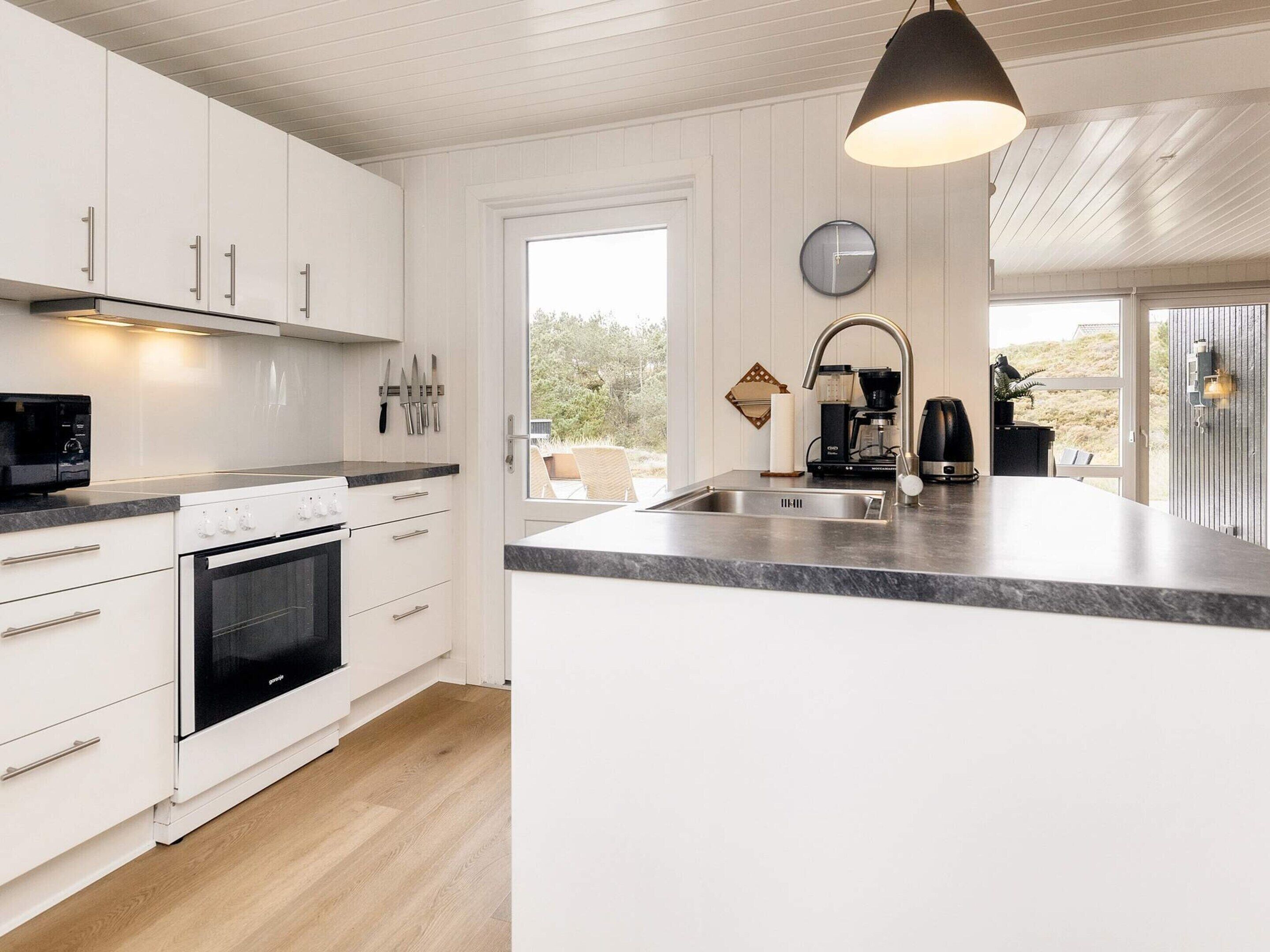 Et moderne køkken med hvide skabe, indbygget ovn, stor ø med vask og hængende lampe.