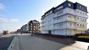 Un edificio residenziale a più piani con balconi, un vialetto lastricato e una spiaggia sullo sfondo.