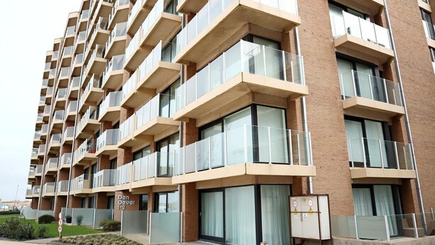 Un immeuble à plusieurs étages doté d’une architecture moderne, avec des balcons pourvus de garde-corps en verre et une façade en briques.
