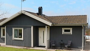 Een gelijkvloers gebouw met een donkere gevel, een pannendak en een schoorsteen. Er bevindt zich een klein terras met twee stoelen en een tafel.