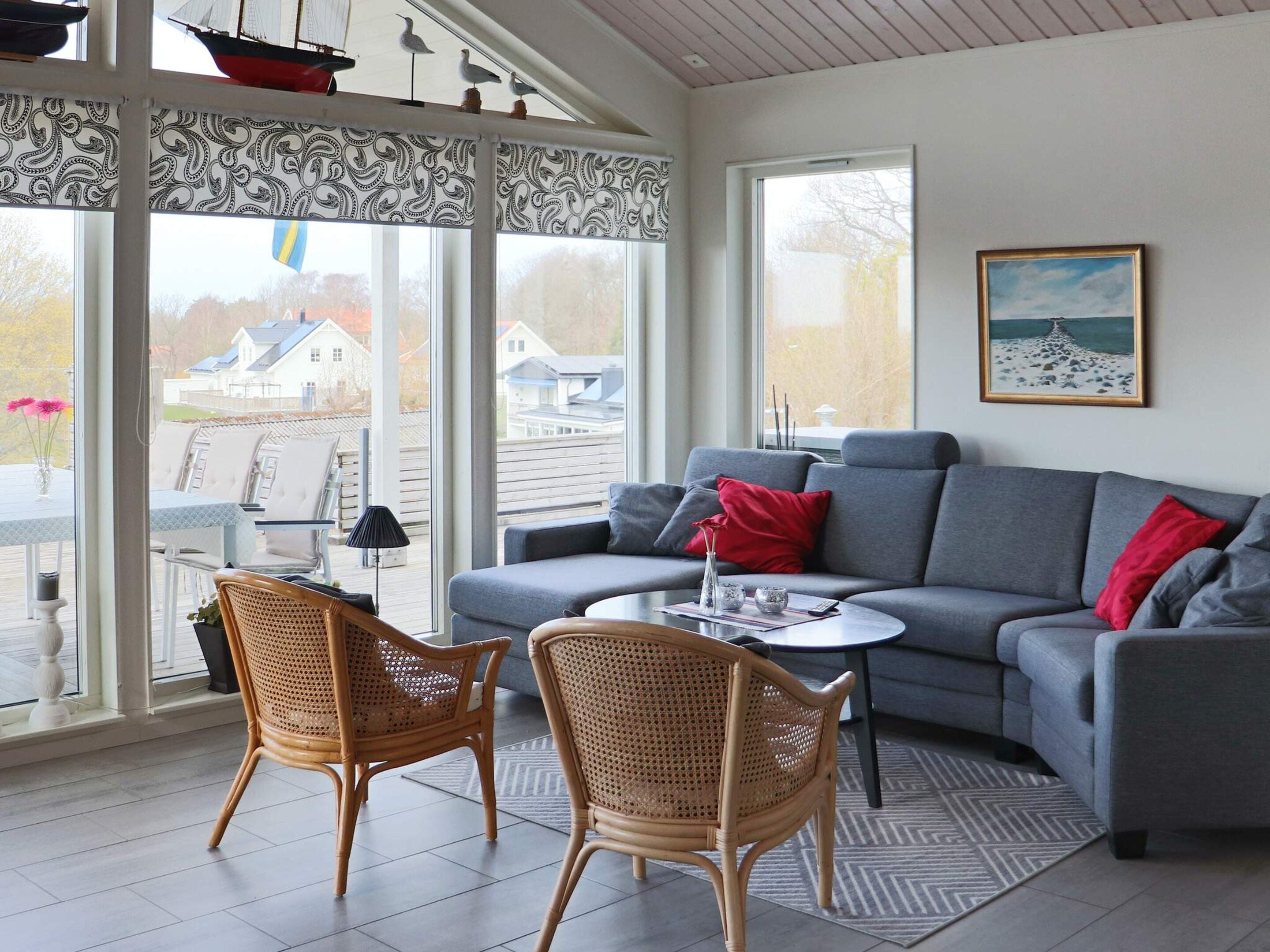 Sala de estar com sofá cinza, duas poltronas de vime, uma mesinha e uma janela grande com cortinas estampadas.