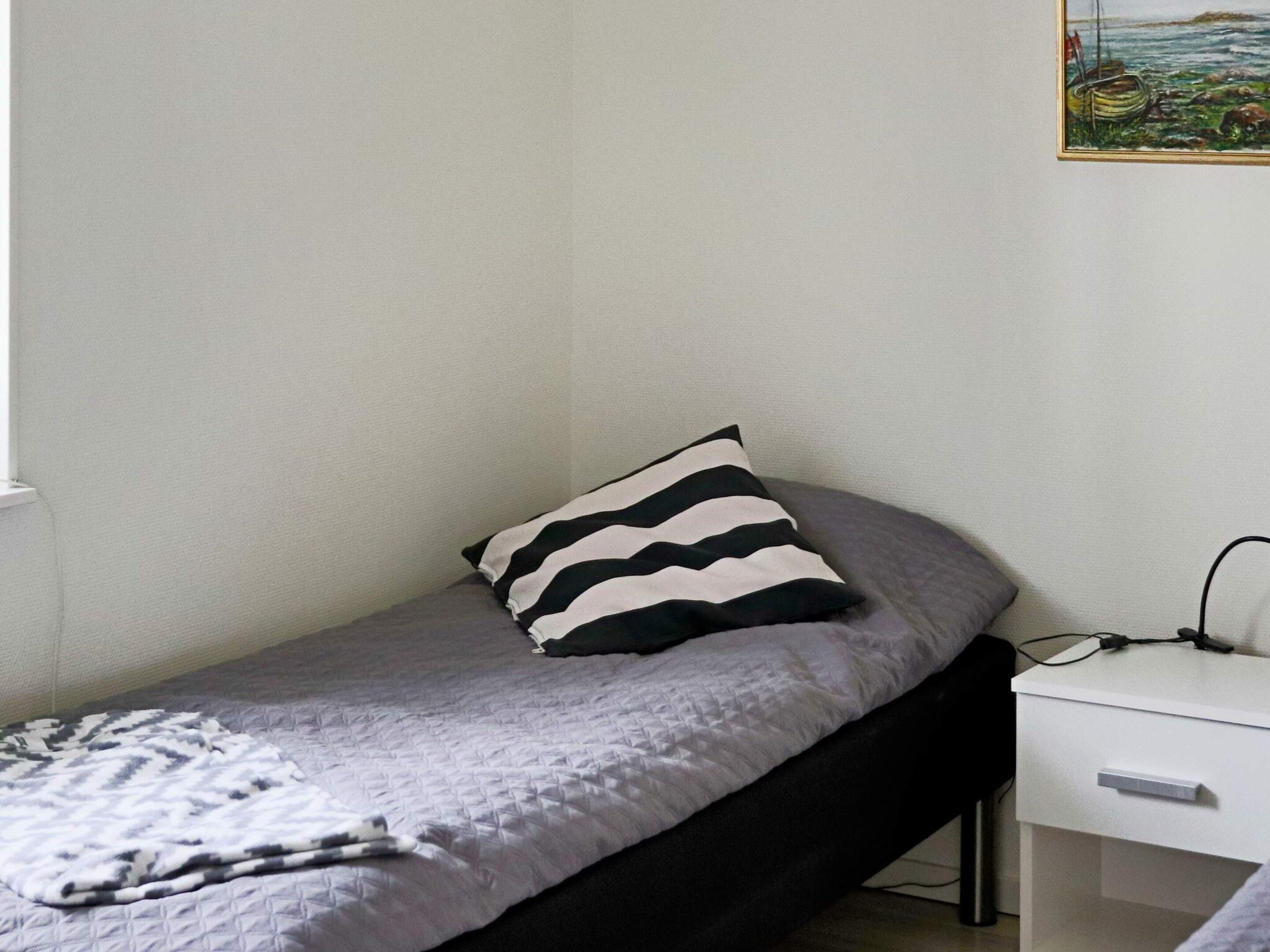 Uma cama de solteiro com travesseiro listrado, uma mesinha de cabeceira com tomada e um quadro pendurado na parede.