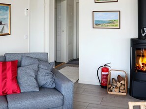 A living room with a grey sofa, red cushion, framed landscape paintings, a black wood-burning stove, and a fire extinguisher. - 5 Star Holiday Home in Steninge (Steninge)