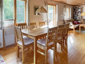 Una mesa de comedor de madera con sillas, una estantería y una lámpara en una habitación con piso de madera y ventanas amplias.