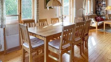 A wooden dining table with chairs, a bookshelf, and a lamp in a room with wooden floors and large windows.