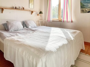 Una cama con cubrecama blanco, estante de madera con objetos decorativos, ventana con cortinas a rayas y un cuadro en la pared.