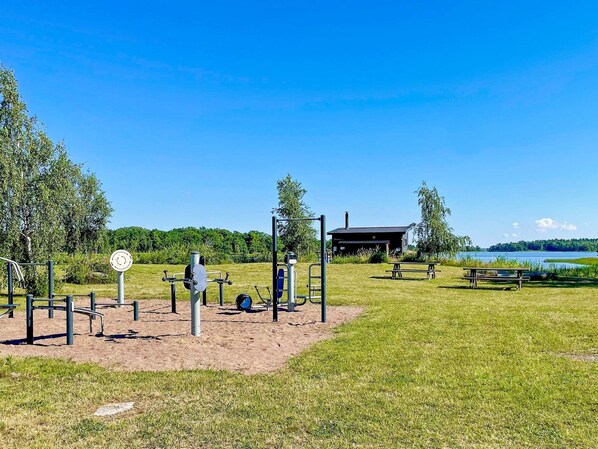 An outdoor gym area with exercise equipment, a covered shelter, and picnic tables by a lake.