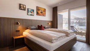Ein modernes Schlafzimmer mit einem großen Bett, einem Nachttisch, einer Bank und Blick auf eine verschneite Landschaft durch ein großes Fenster.