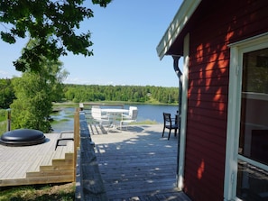 A wooden deck with outdoor furniture overlooking a lake, a red building, and a forested landscape. - 4 Star Holiday Home in Akersberga (Akersberga)