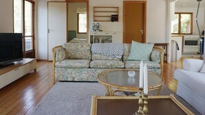 Uma sala de estar com sofá, televisão, mesa redonda com uma vela e cozinha ao fundo.