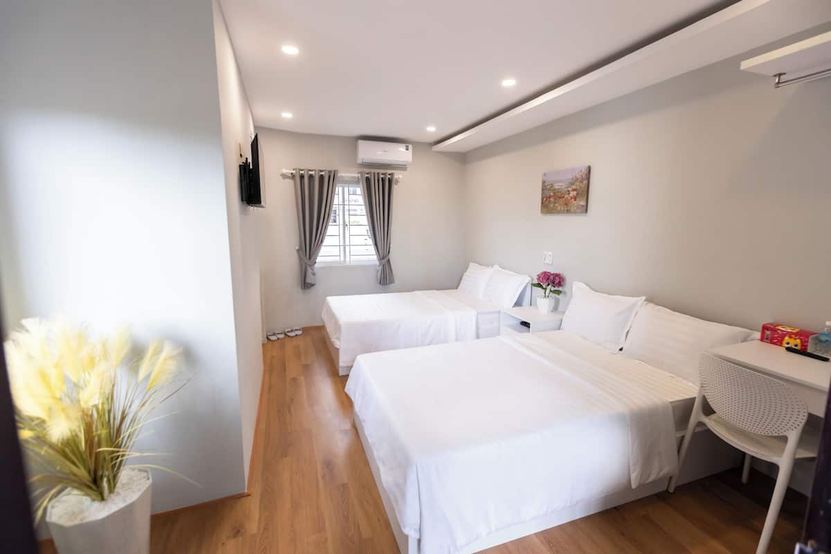 A hotel room with two beds, a desk, a chair, a small table, a vase with flowers, and an air conditioning unit.